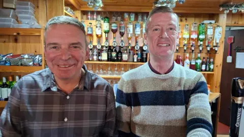 Crown and Anchor Two men with short brown hair standing behind a pub bar and smiling. One had a grey long-sleeved shirt on and the other is wearing a stripy beige, white and blue jumper. He has his arm out as if he is pulling a pint.
