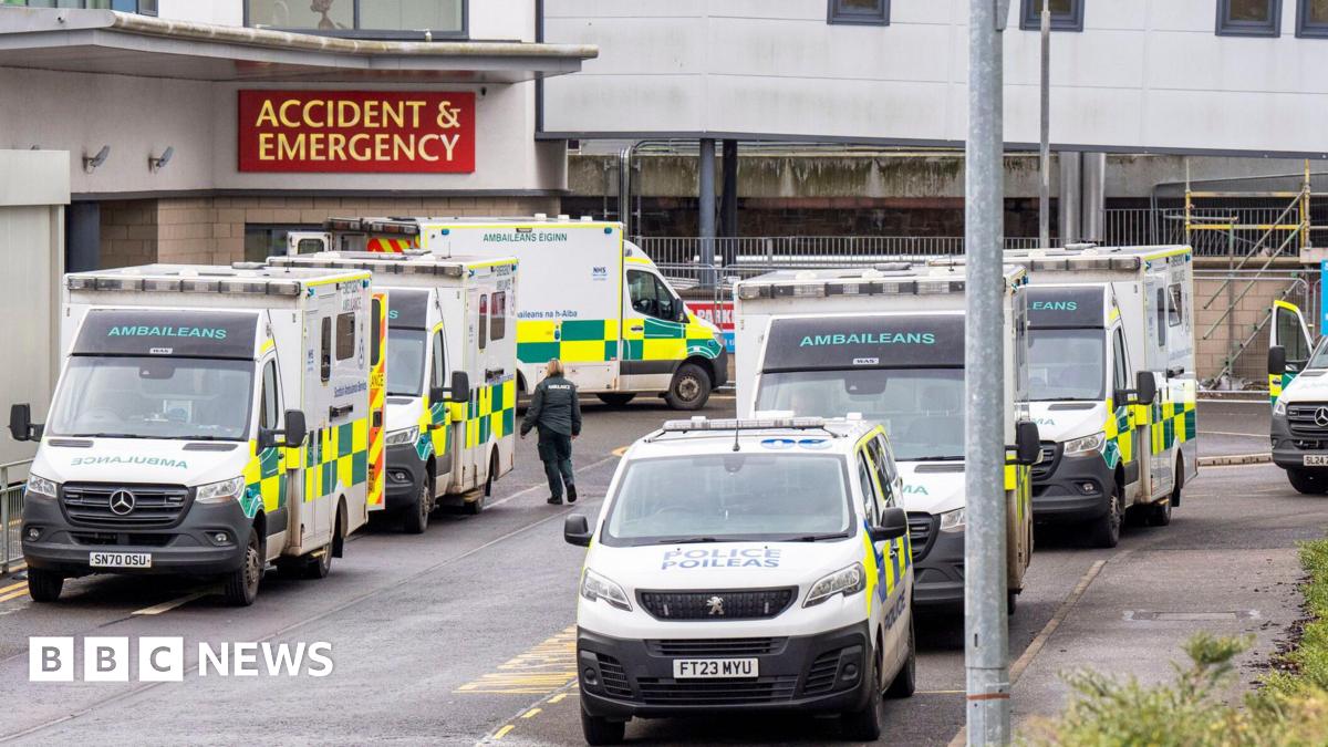An accident and emergency department of a hospital view from the outside. Ambulances and a police van are parked on each side of the road. By the entrance, a red sign has yellow writing which reads ACCIDENT & EMERGENCY
