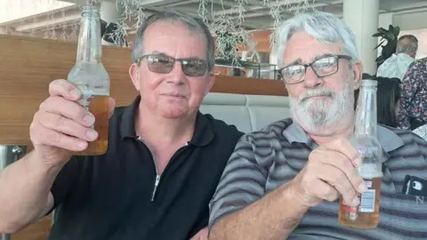 Family photo Two men with glasses raise a bottle of beer each. Both have grey hair, while one has a beard   