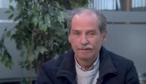 Con Rooney has dark grey hair and is balding on top. He also has a grey moustache. He is wearing a navy winter coat with a light jumper and shirt beneath it.   