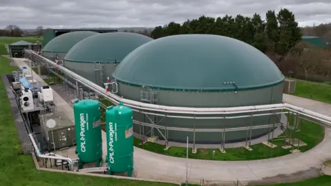 A biogas plant surrounded by grass. It is a large green structure made up of three domes. There are two green cylinders at the front of the image, saying CPL Puragen on them. There are trees in the background.