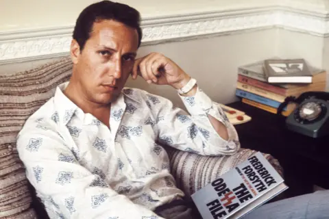 Getty Images Frederick Forsyth is seated on a striped sofa, wearing a patterned button-up shirt and holding an open book titled The Odessa File. Next to the sofa is a small table with a stack of books and a vintage rotary telephone. Behind him are light-coloured walls and decorative moulding.