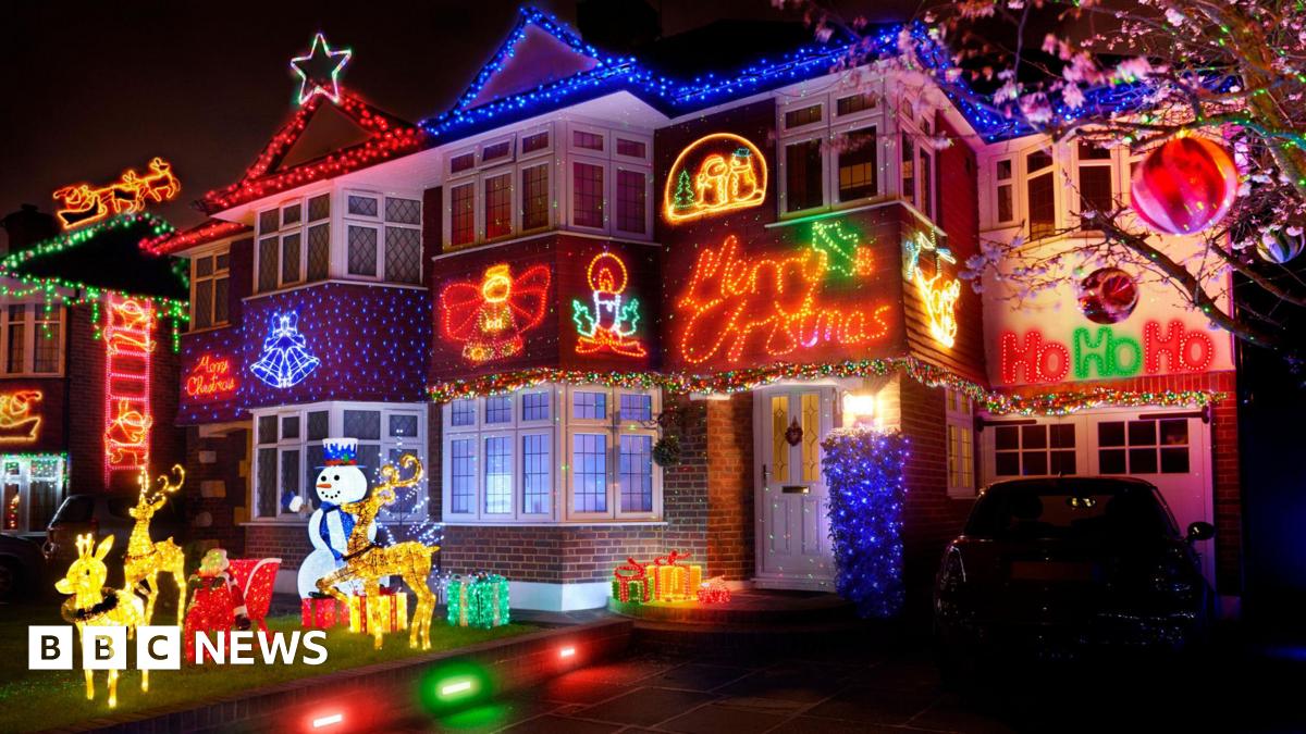 Semi detached house pictured at night with various christmas light decorations including a full santas sleigh and reindeer on the front grass surrounded by light up presents and a huge 'merry christmas' sign lit up above the door.