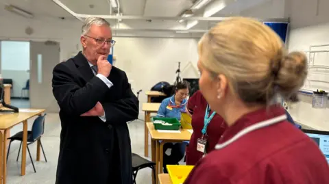 A man in a suit is standing talking to a nurse who is wearing a maroon top. There are other nurses and people standing in the background