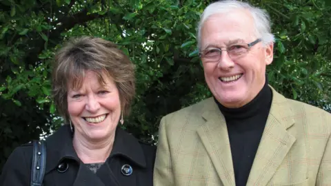 Cancer Research UK Ali and Peter Tomlinson are standing together outside with trees behind them in a close-up photograph. They are both smiling at the camera and looking hapy.