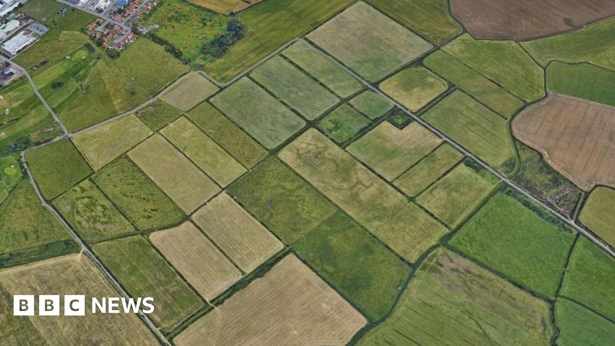 An aerial view of rectangular green fields in Wirral. The land is a patchwork of different greens and browns, with a few buildings - looking like red-roofed newish-build houses and an industrial estate - clumped together in the top left corner.