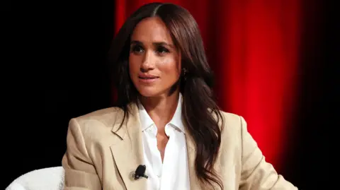 Getty Images Meghan, the Duchess of Sussex sat down on a white chair wearing a beige suit and white shirt