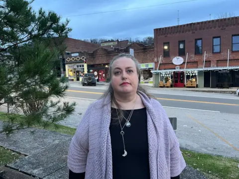 BBC/Ellie House A woman with grey hair and wearing a purple sweater stands in front of a row of shops in the main square in Gainesboro