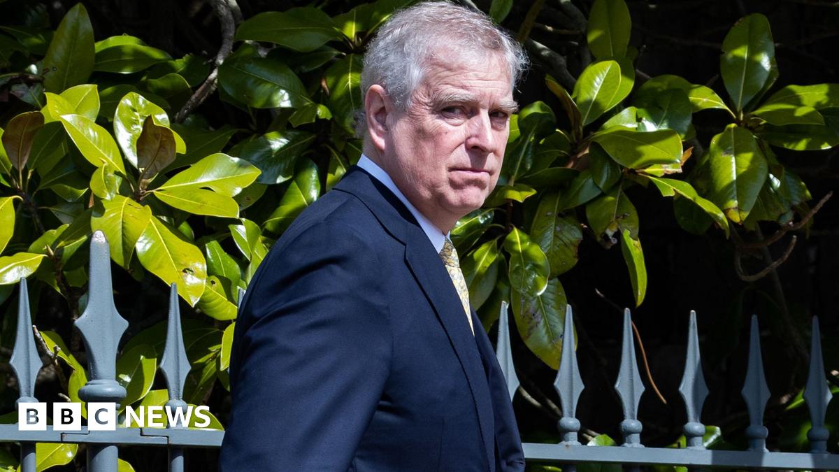 Andrew Mountbatten-Windsor walking outside in a suit looking at the camera on a sunny day. Railings and hedging behind him