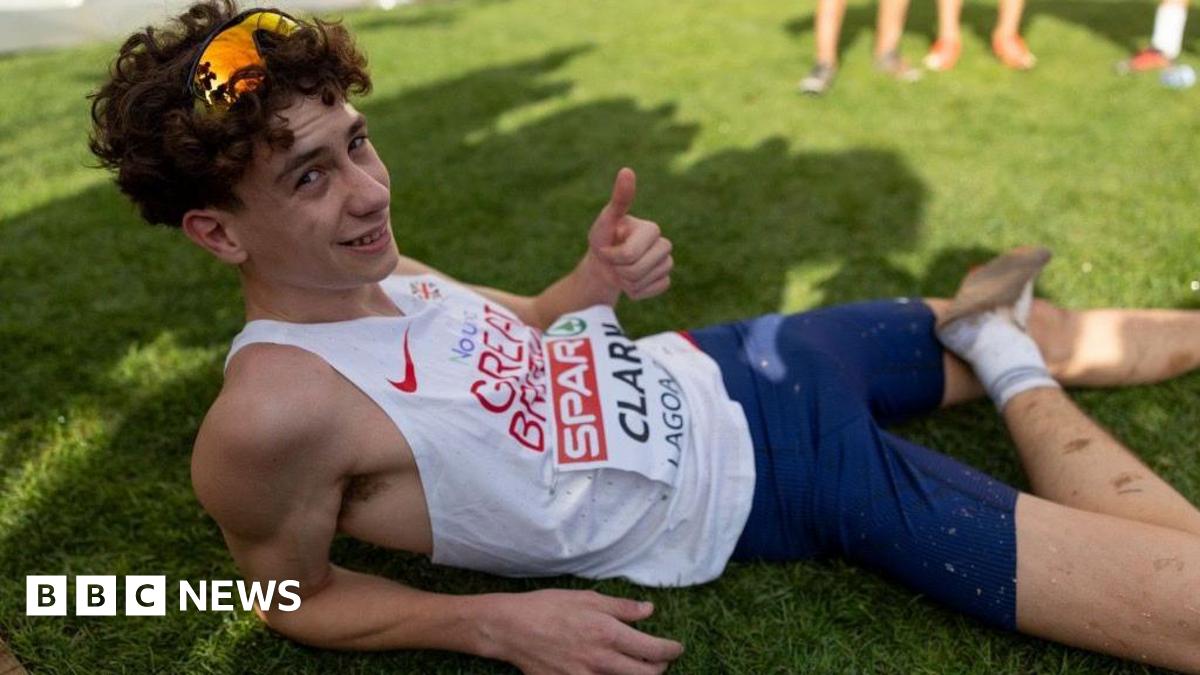 Michael Clark gives the thumbs up while laying on the grass exposing his injured foot. He is in running vest and shorts with his name on his vest bib. He is wearing sunglasses on his head.
