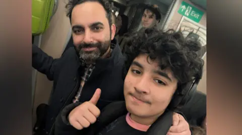 Roy Azoulay A young teenager takes a selfie with his father on board a train. The teenager has a black jacket and black curly hair and his father Roy has black curly hair and a beard. Other commuters can be seen standing behind them