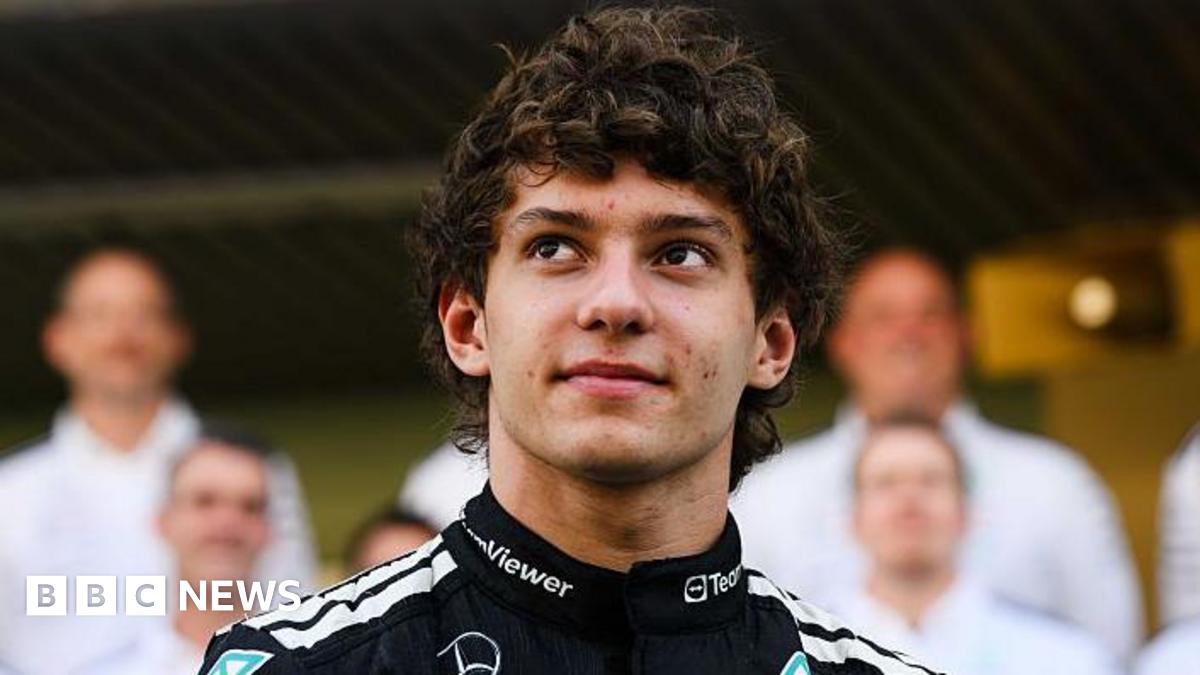 Kimi Antonelli as a racing driver looking up, with a black racing top, with people behind him, blurred. He has short dark curly hair.