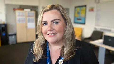 Dan Nelson / BBC A woman with blue eyes and shoulder length blonde hair is smiling at the camera. She's wearing a staff lanyard and a black jacket. She's standing in the middle of a school office. 