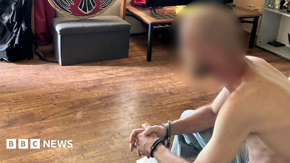 Two men, both with their backs to the camera. One man has his hands cuffed behind him.