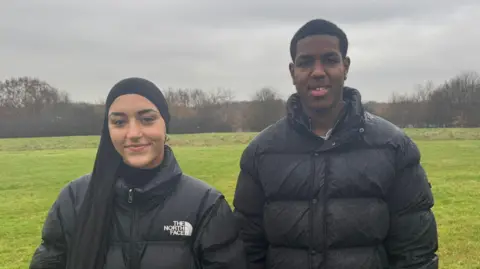 Zainab is wearing a black hijab and a black North Face coat. Jaiden is wearing a black puffer coat. The are stood side-by-side in a field with trees in the distance. 