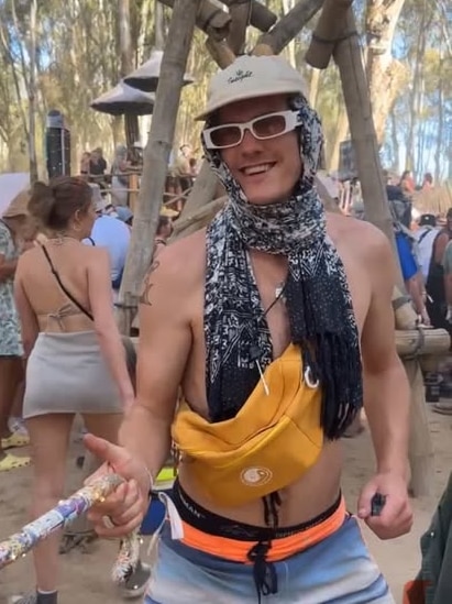 A young man at a music festival holds a walking stick. He's wearing sunglasses, a hat and scarf.