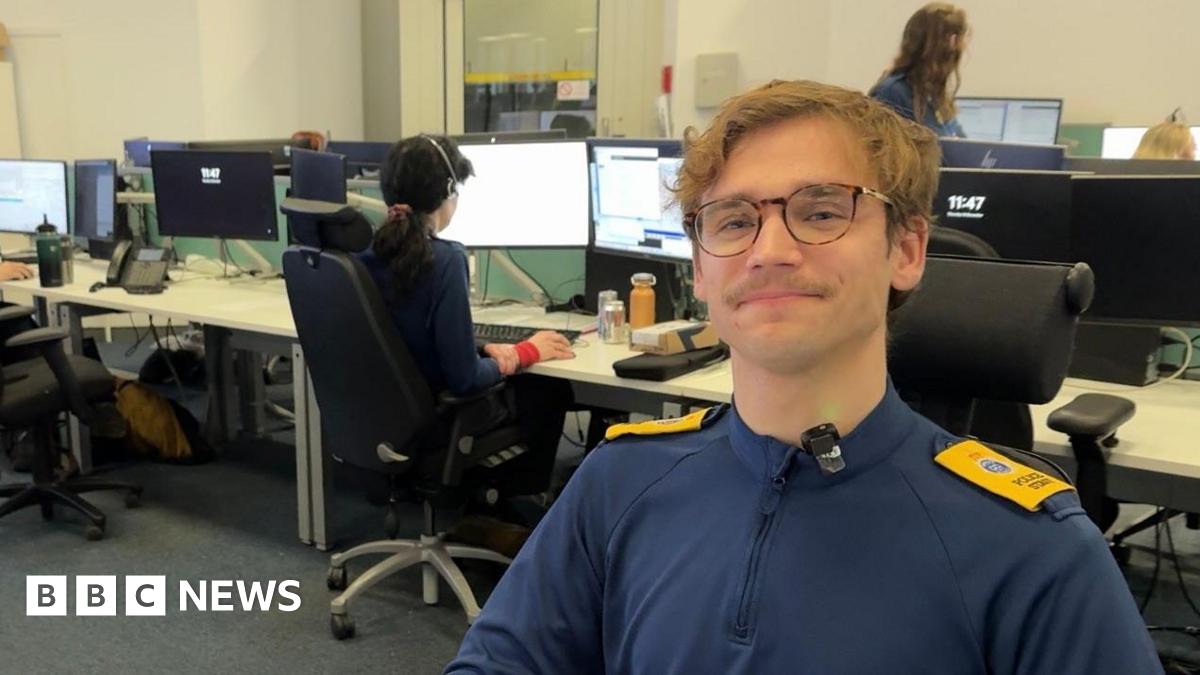 A man wearing a navy blue top with yellow shoulder pads. He is sat in an office with people and computers in the background.