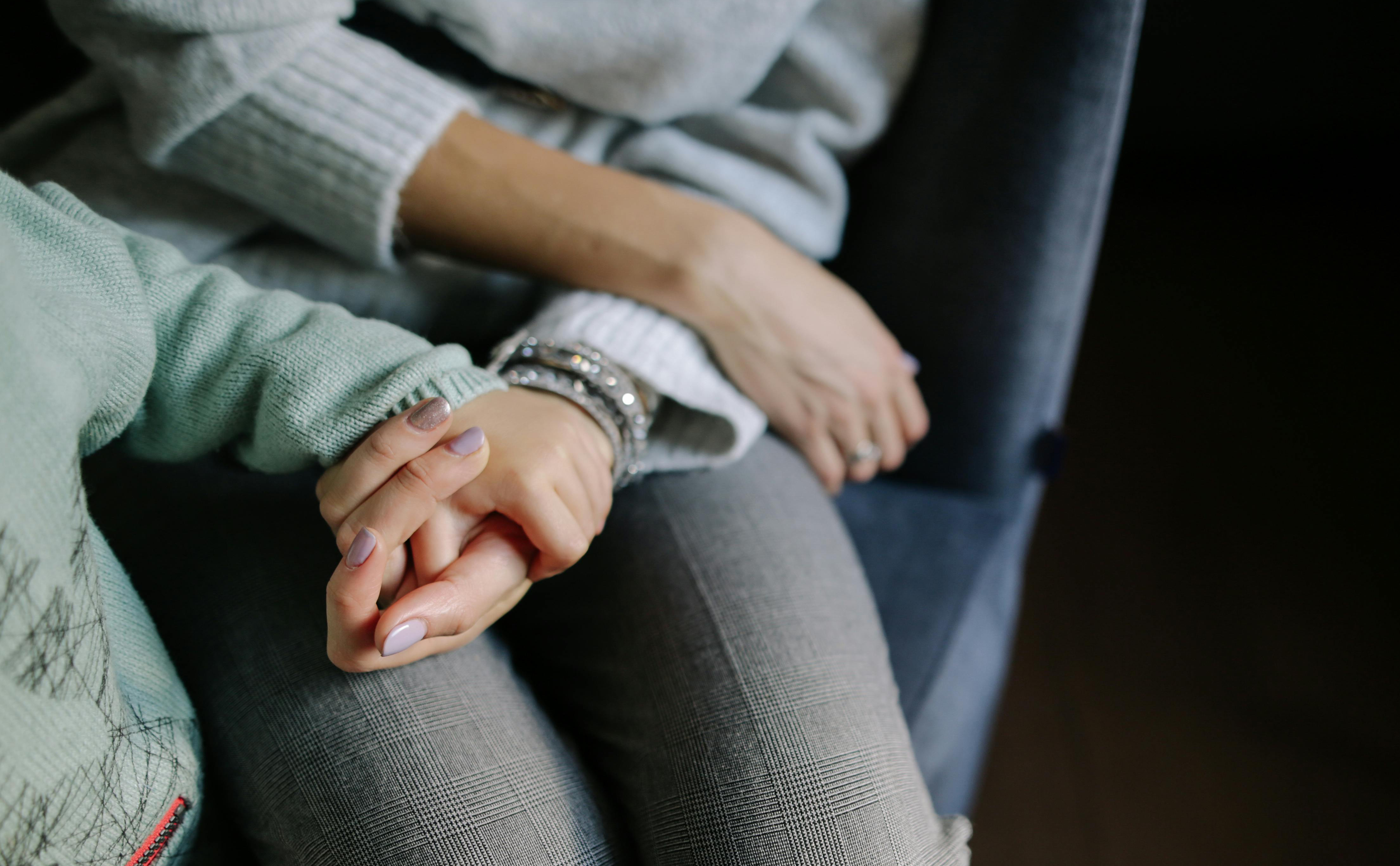 A woman’s hand cupped upwards holding a toddler’s hand with both rested on the woman’s leg.