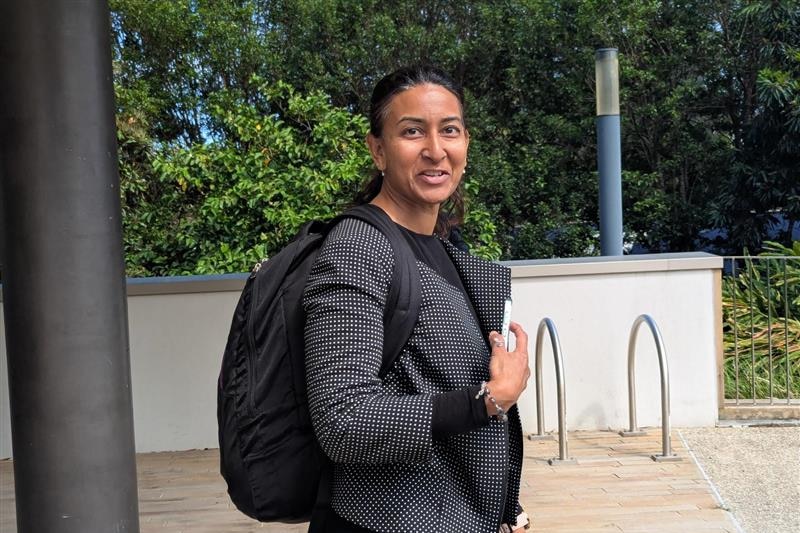 A dark-haired woman, formally dressed and wearing a backpack, stands outside a court building.