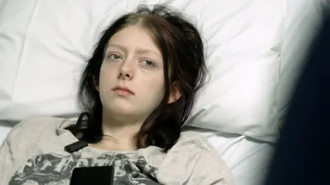 BBC Paige who is 19 is lying on a hospital bed resting her head on a pillow. She looks ill. She has dark hair and is wearing a T-shirt.
