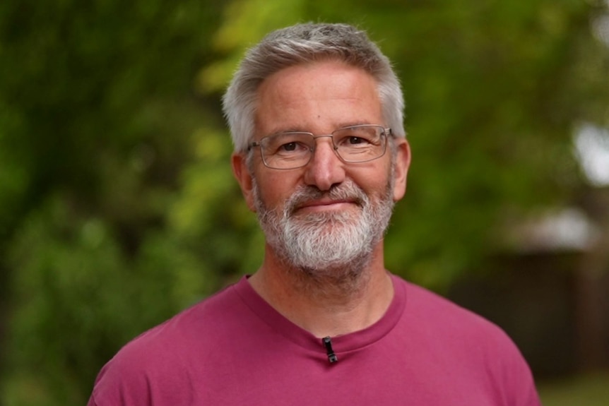 A t-shirt clad, bespectacled, middle-aged man with greying hair and beard smiles at the camera. 
