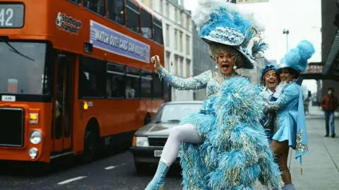 Getty Images Stanley Baxter stands at a bus stop, dressed as a panto dame in a blue dress and large hat. He is sticking his leg out into the road comically, as a large red bus with a Strathclyde Transport sign drives by. Two other cast members stand behind him, laughing.