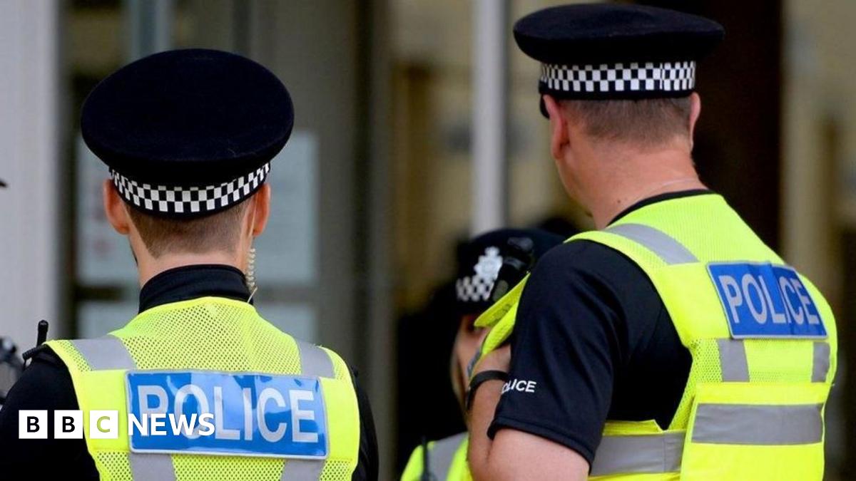 Three police officers pictured from the back wearing black polo shirts, yellow hi-vis vests with the word POLICE, and black caps with black and white chequered brims.