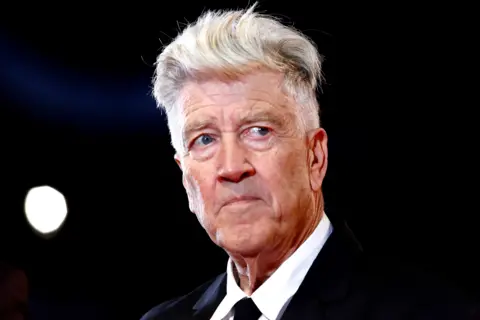 Getty Images David Lynch has light-coloured, voluminous hair styled upward and is wearing a formal black suit jacket with a crisp white dress shirt and a black tie. The background is dark, with a bright circular light source visible on the left side, creating a dramatic contrast and highlighting the hair and upper part of the outfit.