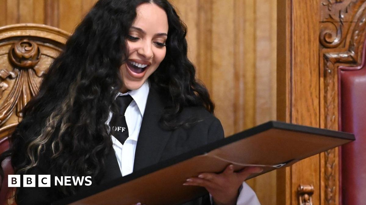 Jade Thirlwall looking down at a picture she has been presented with from South Tyneside Council. She has long black hair and is wearing a black blazer and tie with a white shirt. She has her mouth open wide in a smile.