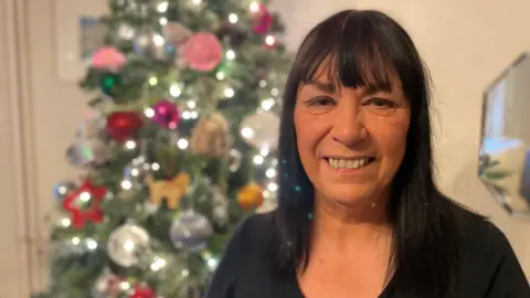 BBC A woman with shoulder length dark hair, wearing a black top standing in front of a Christmas tree.