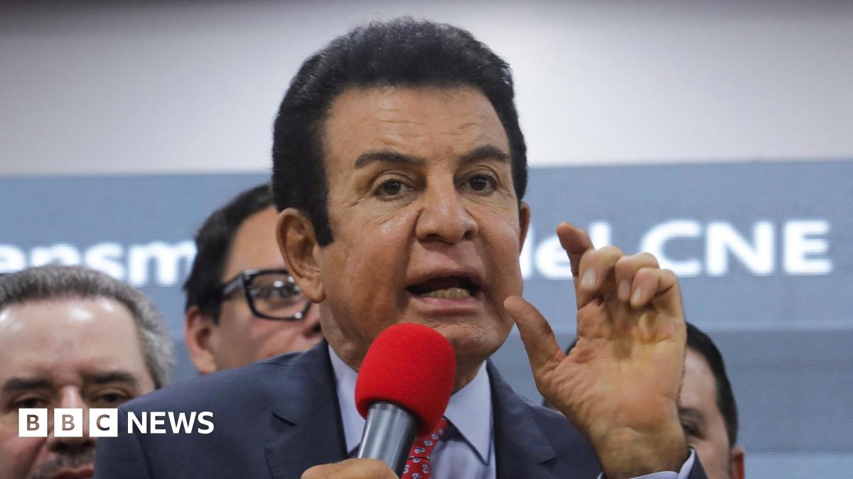 Presidential candidate Salvador Nasralla of Honduras' Liberal Party (PLH) gestures during a press conference. He wears a navy suit, blue short and red patterned tie. He is speaking into a bright red microphone