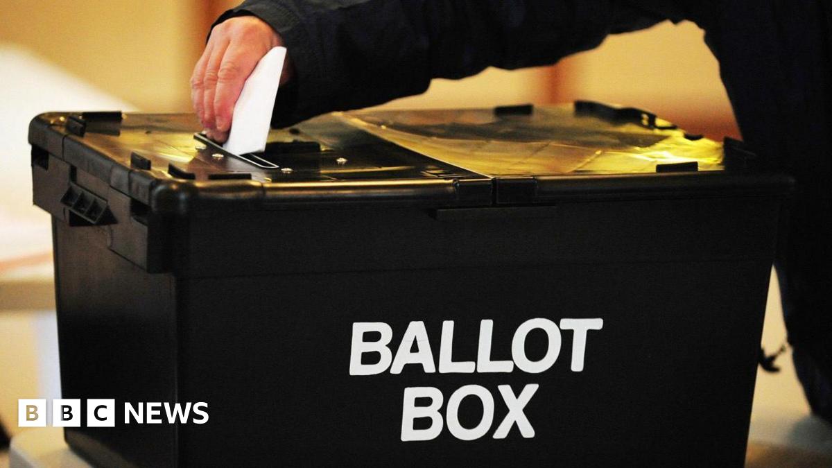 The hand of an unknown person placing a voting slip in a black ballot box with the words marked clearly on the side in white capital letters.