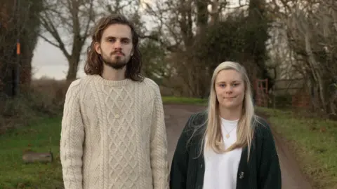 Oliver and Abbie Stone Houhgton. Oliver is wearing a cream woollen jumper, Abbie is wearing a white t-shirt and dark green cardigan. Behind them are trees and a path