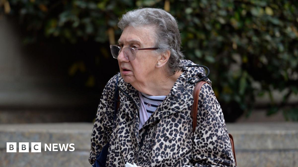 Patricia Robertson, a 77-year-old woman with short grey hair and clear-rimmed glasses, is walking right to left. She is wearing a leopard skin hooded jacket and a  light and dark blue striped shirt underneath. She has a brown leather bag slung over her shoulder.