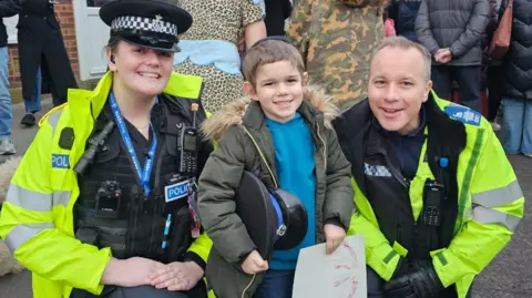 Chabad Lubavitch Bricket Wood A female and male police officer crouch down either side of a boy, all smiling at the camera. He holds a piece of paper with colouring-in, and the hat of one officer. The officers wear high vis neon coats over their black police vests, which have phones and body cams on. 