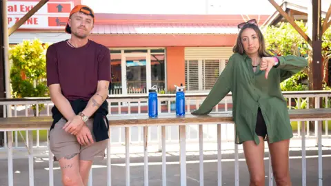 StudioLambert/BBC Broadcaster Roman Kemp and his sister, singer-songwriter, Harleymoon leaning against a wall whilst waiting for transport. Harleymoon signals a thumbs down with her hand.