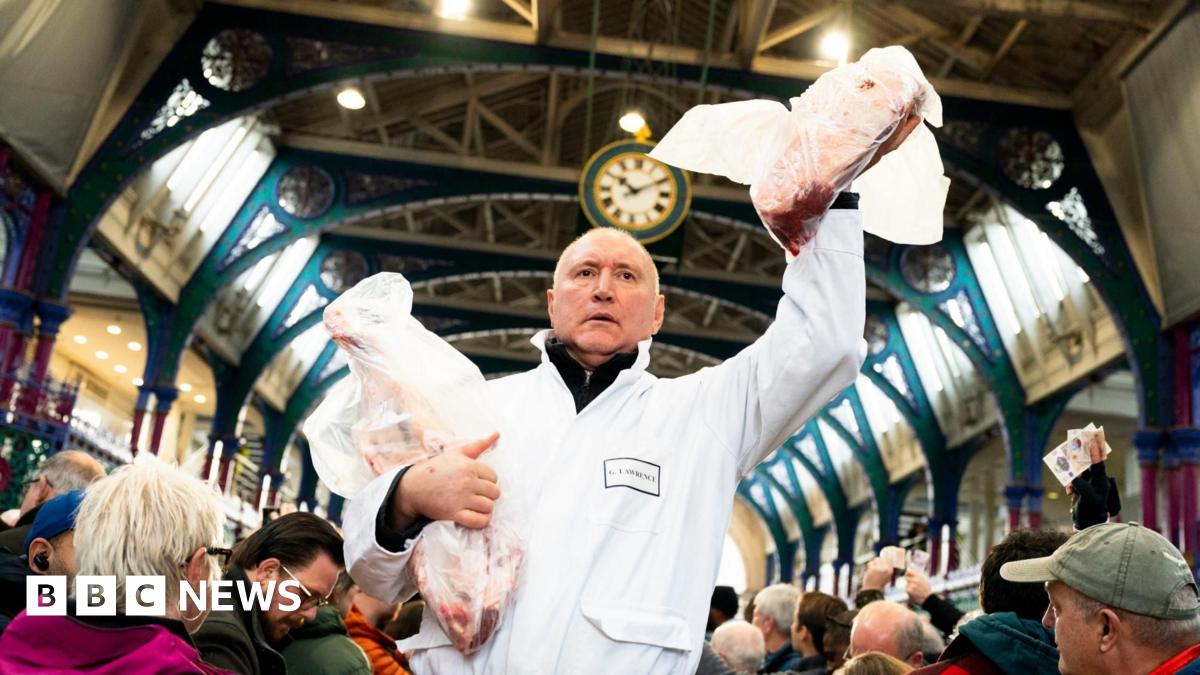 A trader wearing a white overall holds to joints of meat - one of which in the air - while people stand around him in Smithfield meat market