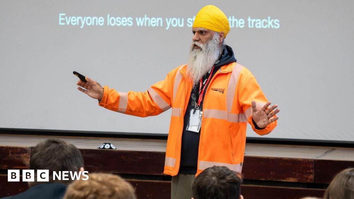 A man with a long white beard and moustache standing in front of children, talking. He is wearing a yellow head scarf, red lanyard and orange hi-vis jacket