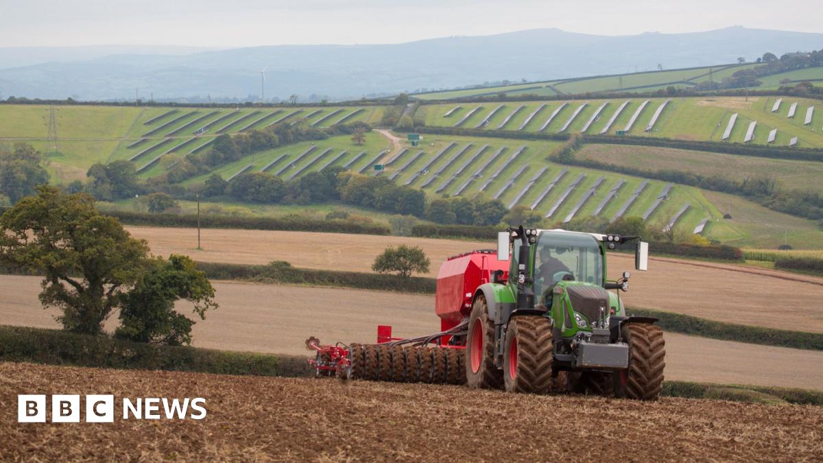 A green and red tractor is pulling a cultivator and drill across a muddy field. On the hills in the background lie solar panels. A light mist sits in the sky.