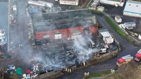 Fife Jammer Locations Aerial view of the smouldering aftermath of a fire at a yard next to a harbout. Two fire engines and several firefighters can be seen in the image.