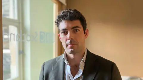 A dark haired man with a grey suit jacket and white shirt looks at the camera