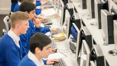 Getty Images Three boys wearing blue school jumpers and white shirts sit in front of computers in a classroom. They are looking straight ahead at their screens