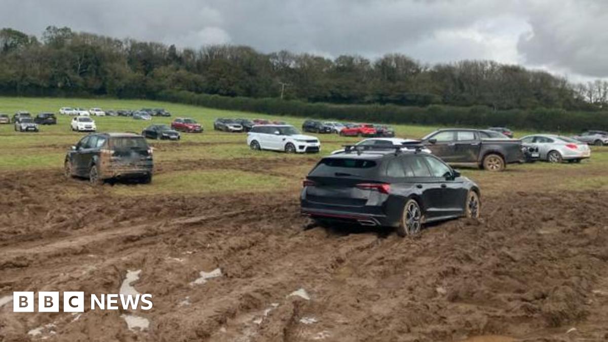 A field covered in wet mud with dep track marks from car tyres. Some of the cars in the foreground are covered in mud while others are parked in the distance.