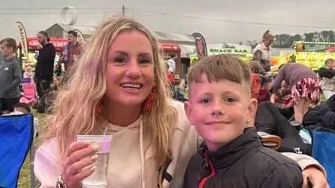 Family photo Nicola and Zac at a festival. They are both smiling at the camera. Nicola is hold a plastic cup with her arm around zAC. 