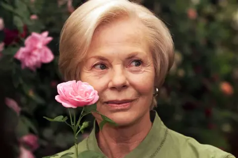 Getty Images The Duchess of Kent, wearing a light green collared garment, is holding a pink rose close to the face. The background is filled with lush greenery and several blooming pink roses, creating a garden-like setting. The lighting is soft, highlighting the flower and the surrounding foliage.