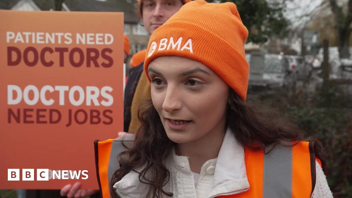 Dr Heather Gunn has long, curly brown hair under her orange BMA beanie hat. She is wearing an orange fluorescent jacket and standing on the picket line, next to a sign that reads "Patients need doctors. Doctors need jobs."
