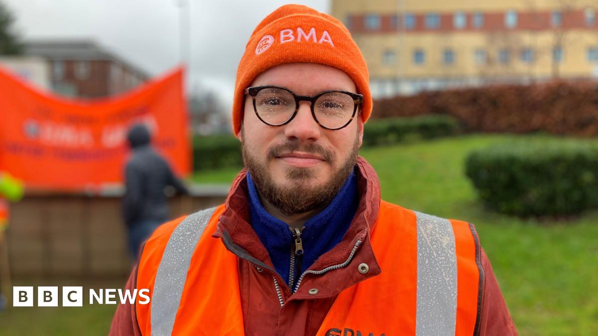 A man with brown facial hair and black glasses with rain on the lenses. He is wearing an orange beanie with white writing that says "BMA". He is wearing a red waterproof jacket and an orange and silver hi-vis jacket on top.