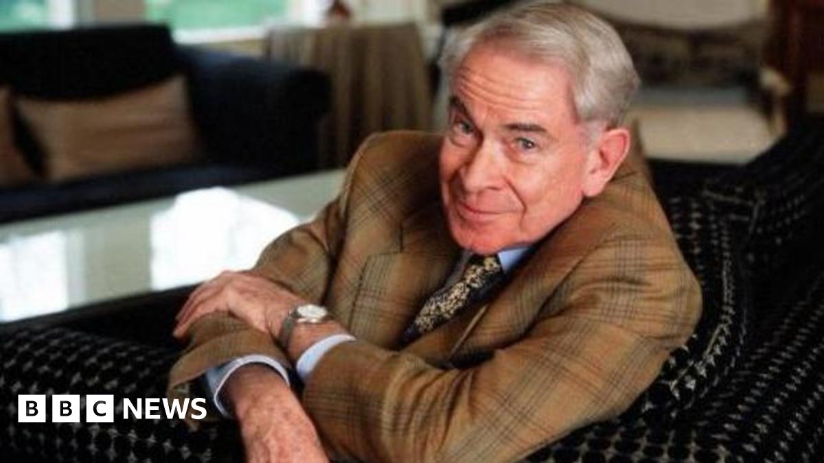Stanley Baxter is photographed in later life.  He is looking at the camera wearing a suit and he has his arms crossed.