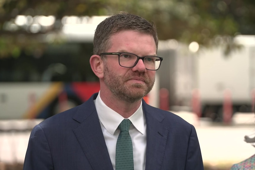 Chris Picton wears glasses and a suit as he stands in a park with traffic behind him.