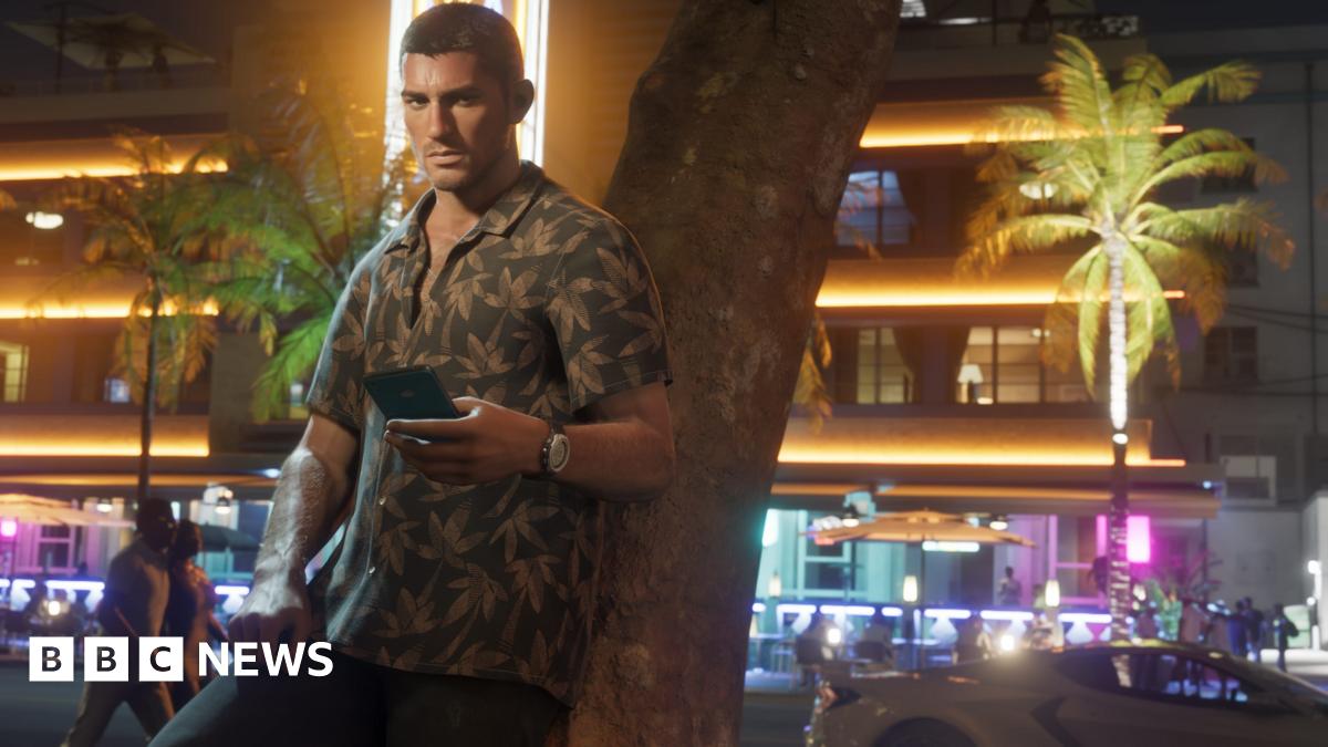 Screenshot shows Jason, a main character from GTA 6, wearing a green Hawaiian shirt with palm tree pattern, leaning against a tree as he holds a phone in one hand. Behind him are white seafront buildings bathed in a mix of yellow and purple lights.